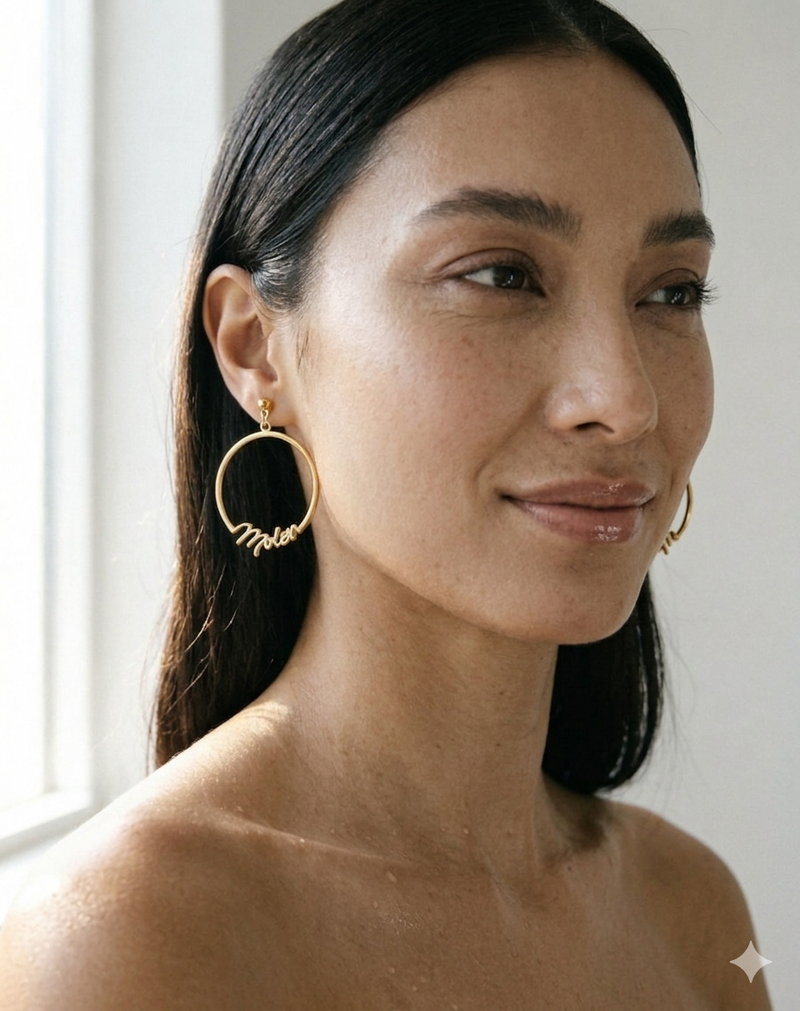 A close-up of a woman wearing a pair of gold hoop earrings with cursive script text MOIEN on the front, set against a bright, softly lit background