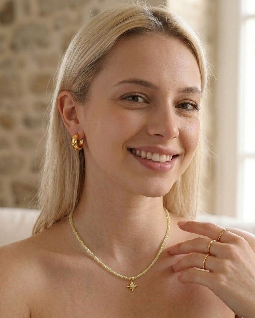 Woman wearing gold jewelry including a necklace, earrings, and rings against a stone wall background.