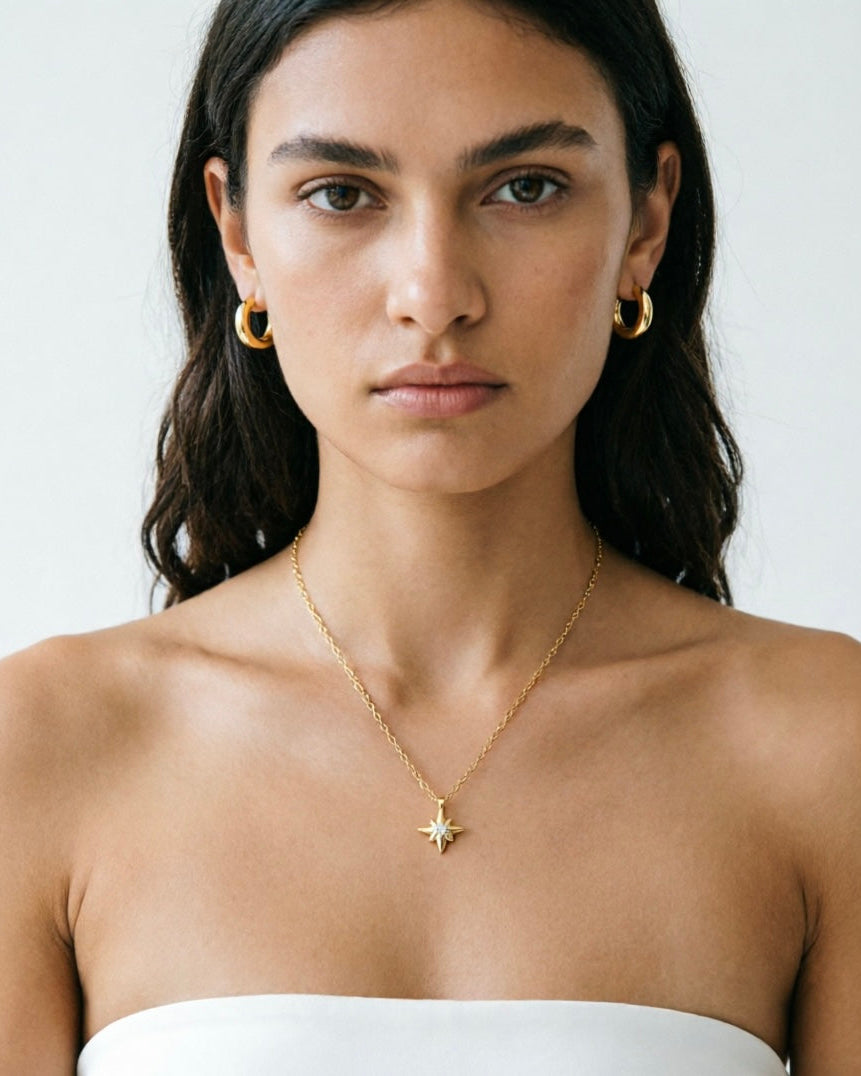 A close-up of a model wearing a delicate gold necklace with a starburst pendant and matching gold hoop earrings, paired with a white strapless top against a light gray background.