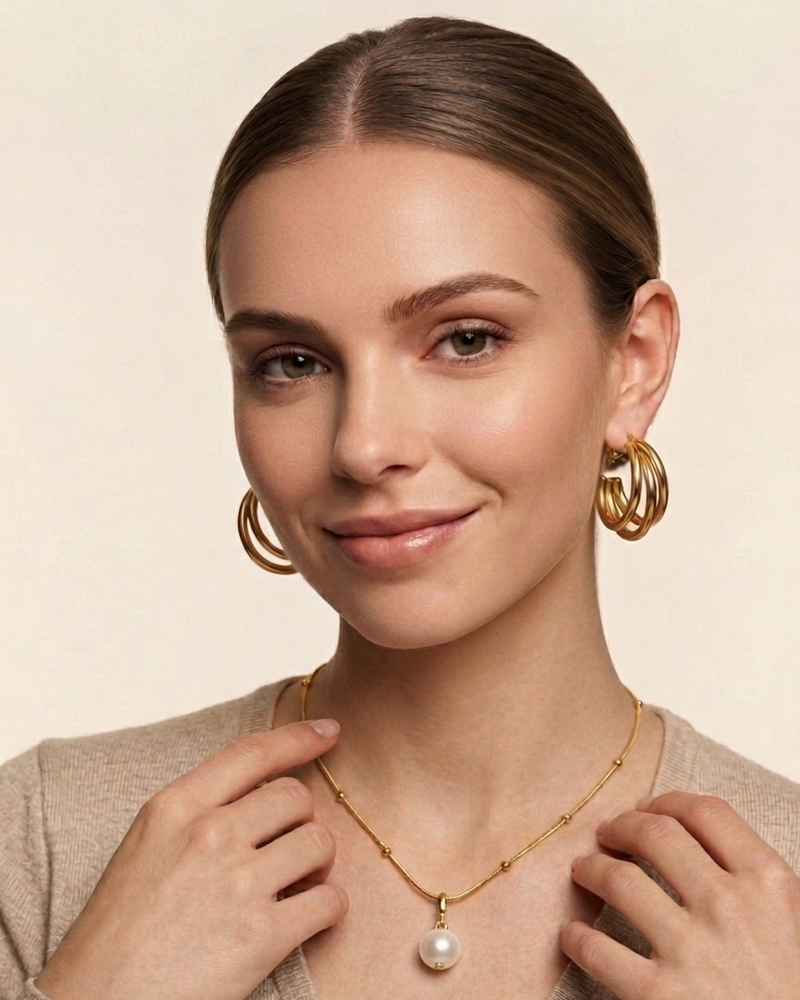 A woman with her hair pulled back wears a delicate gold necklace with a white pearl pendant and matching gold hoop earrings,