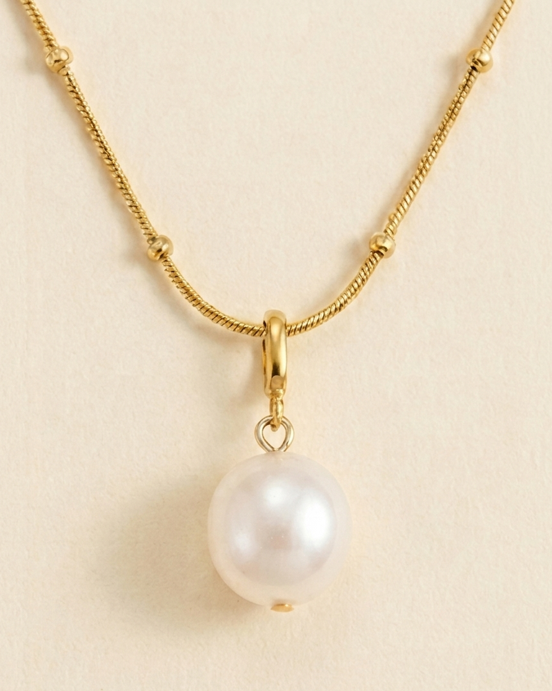 A gold chain necklace featuring a single round white pearl pendant, displayed against a light beige background.