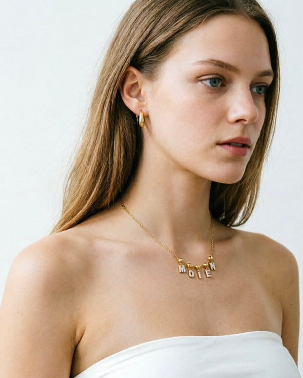 A woman with long light brown hair wearing a delicate gold necklace with the word 'MOIEN' spelled out in small letters, paired with a white strapless top against a plain light gray background.