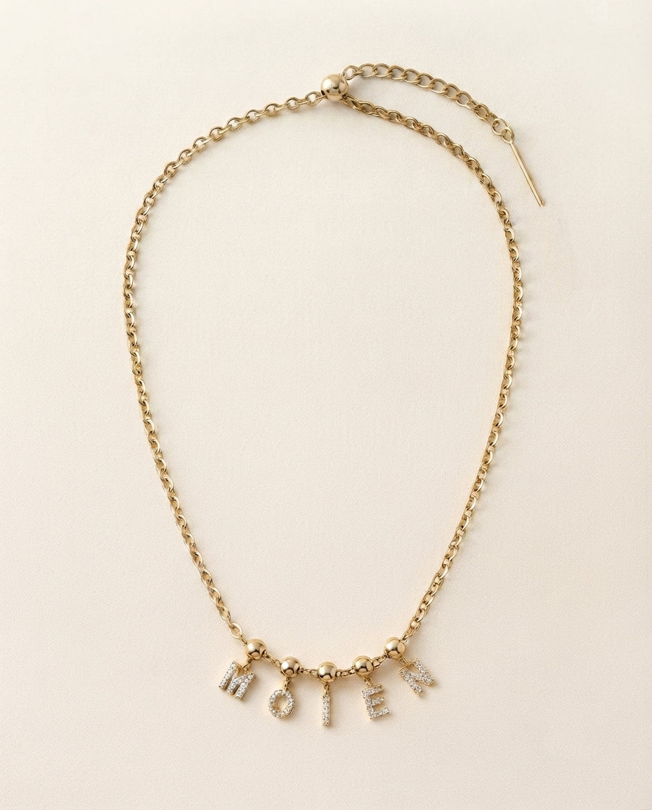 A gold chain necklace with a delicate drop pendant spelling out 'MOIEN' in small letter charms, displayed on a light cream background.