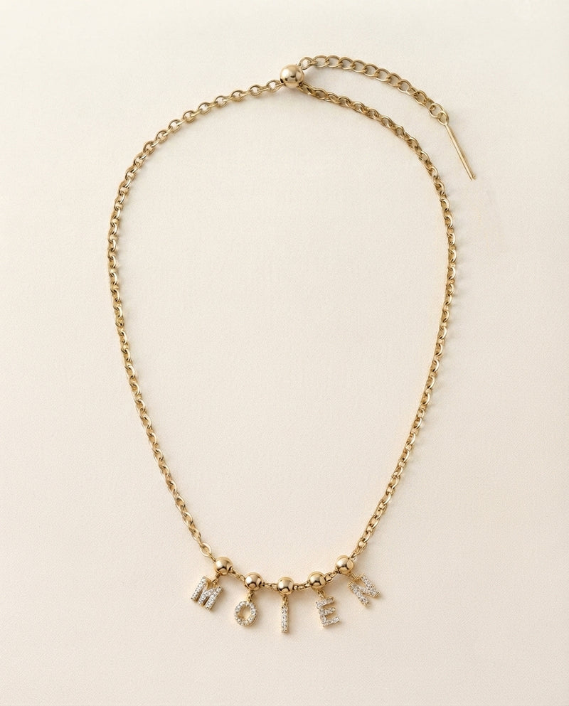 A gold chain necklace with a delicate drop pendant spelling out 'MOIEN' in small letter charms, displayed on a light cream background.