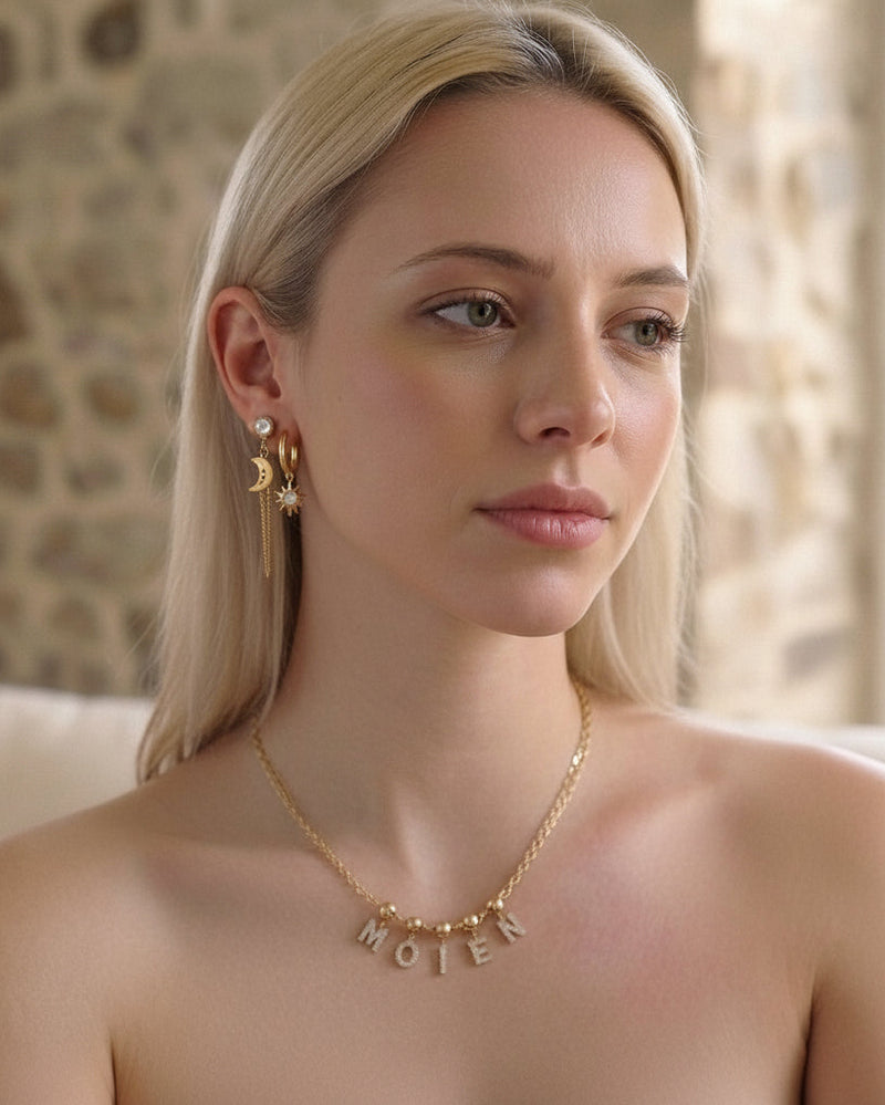 A blonde woman wearing a gold chain necklace with the word 'MOIEN' spelled out in letter charms, shown in a close-up portrait against a softly blurred stone wall background.